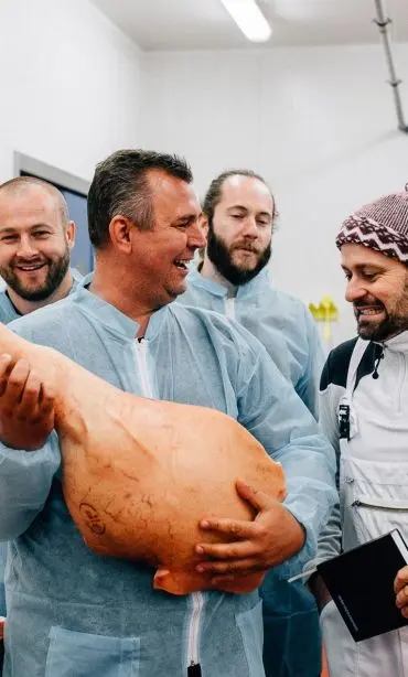 Skupina mužů v ochranných oblecích si prohlíží velkou vepřovou kýtu a směje se.